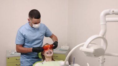 Ağız retraktörü, diş aletleri, diş muayenesi. Mavi önlüklü ve maskeli diş hekimi, diş sandalyesinde otururken ağız retraktörü takan gülümseyen hastaya koruyucu göz giysisi ayarlıyor..