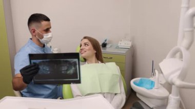 Dişçi önlüğü, diş muayenesi, diş sağlığı. Mavi önlük ve maske giyen dişçi diş röntgenini dişçi koltuğunda gülümseyen hastaya gösteriyor..
