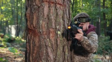 Modern tüfek, taktik pozisyon, düşman çatışması. Orman ortamında, kafasında işaretle kamuflaj kıyafeti giymiş bir adam ağaca yaslanırken lazer tabancasıyla nişan alır.
