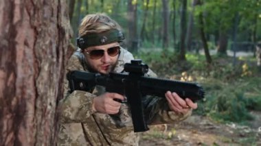 Düşmanı vurur, ağaç duruşu, düşman savaşı. Kafasında işaretle kamuflaj kıyafeti giymiş bir adam ağaca yaslanmış, lazer silahı doğrultuyor ve ormanda rakibini vuruyor..