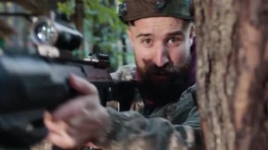 Orman çevresi, odaklanmış hedef, açık hava oyunu. Ormanda, kafasında işaretle kamuflaj kıyafeti giymiş bir adam ağaca yaslanır, kasıtlı olarak lazer tabancasını hedef alır ve rakibine ateş eder.