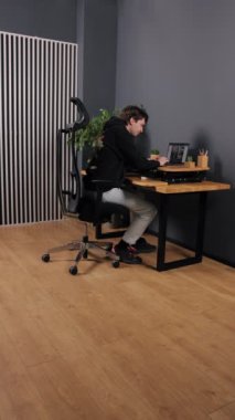Laptop görevleri, dijital görevler, ahşap ofis. Dikey videoda, siyah kapüşonlu adam, siyah ofis sandalyesi ve çeşitli aksesuarlarıyla ahşap masada dizüstü bilgisayarda çalışıyor..