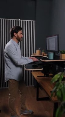 Masa ayakta, mavi bitkiler, çalışan monitör. Açık mavi gömlekli adam modern ofiste monitörle ayakta duruyor. Ahşap zemin ve bitkiler, dikey video..