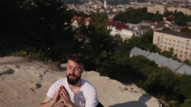 Yoga refahı, beyaz tişört, yoga gücü. Genç adam yogayı tepe örtüsünde yapıyor. Doğal ortamdaki konsantrasyon ve istikrarın altını çiziyor. Arkasında çarpıcı bir şehir manzarası var..