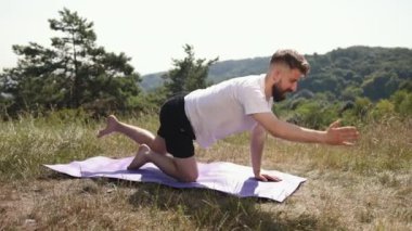 Denge pozu, yoga ahengi, doğayı birleştirme. Siyah şort ve beyaz tişört giyen genç adam, doğa dolu çimenlik alanda mor paspasın üzerinde yoga dengesi kuruyor..
