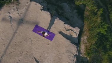 Hava manzarası, yoga pratiği, poz pratiği. Üst görüş, mor minder üzerinde yoga yapan hava manzaralı kişi kayalık yüzeyde yatıyor, şahıs dengeli bir şekilde günlük atletik kıyafetler giyiyor.