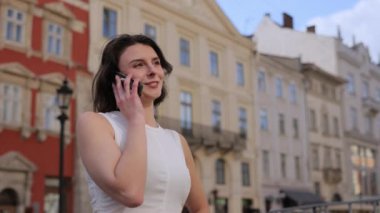 Kadın araması, şehir merkezi, tarihi binalar. Beyaz gömlekli kadın telefonu kulağına tutarken gülümsüyor, tarihi binalarla şehirde duruyor..