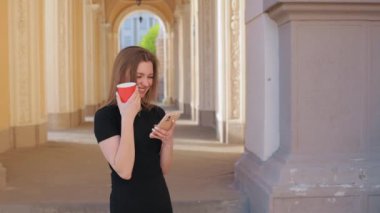 Cep telefonu, kırmızı bardak, cep telefonu kullanımı. Siyah elbiseli kadın akıllı telefonunu kullanıyor ve elinde kırmızı kahve fincanı, kemer yolunda süslenmiş sütunlarla duruyor..