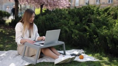 Bağlı kalmak, serbest çalışmak, Doğa Ofisi. Arkaplanda çiçek açan sakura ağacının ortasında, kadın kendini dışarıda bilgisayarından video aracılığıyla iletişim kurarken buluyor..