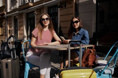 Açık hava kafe, gülümseyen kadınlar, güneşli kafe. İki genç kadın açık kafede güneşli bir günün tadını çıkarıyorlar, bavullarıyla oturup gülümsüyorlar, rahat seyahat anlarını yakalıyorlar..