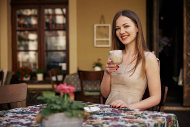 Elinde kahve fincanı, açık kahve, gülümseyen kadın. Genç bir kadın kahve bardağını açık kafede tutarken gülümsüyor, sıcak ve rahat bir atmosfer yaratıyor..