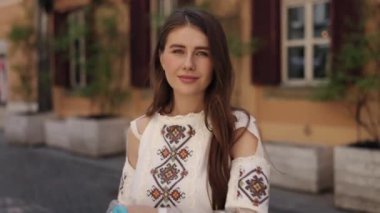 Sokak arkaplanı, bayrak sallama, geleneksel elbise. Antik şehir manzarasında, genç kadın gururla mavi ve sarı bayrak sallıyor, işlemeli bluzu kültürel gururunu yansıtıyor..