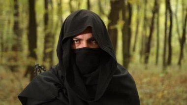 Kapüşonlu figür, karanlık pelerin, gizemli figür. Siyah kapüşonlu ve maskeli, ormanlık alanda yoğun bir şekilde bakan gizemli kişi..
