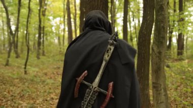 Gizlenmiş figür, sırtında kılıç, Woodland sahnesi. Arkadan bakıldığında siyah bir figür ormanda yürüyor, sırtında yıpranmış bir kılıç taşıyor..