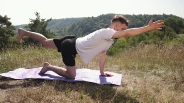 Doğa bağlantısı, yoga sükuneti, konsantrasyon dengesi. Doğayla iç içe olan beyaz tişörtlü ve siyah şortlu genç adam, çimenli alanda mor yoga minderinde farkındalık egzersizi yapıyor..