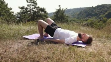 Yoga yolculuğu, yoga meditasyonu, barışçıl doğa. Çimen tarlasında mor hasırda, beyaz tişörtlü ve siyah şortlu genç adam farkındalık, denge ve doğal çevreyi kucaklama pratiği yapıyor..