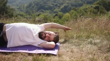 Fitness dengesi, denge pozisyonu, yoga dengesi. Beyaz tişört ve siyah şort giyen genç adam, doğanın kuşattığı çimenli alanda mor paspas üzerinde yoga yaparken dengeye odaklanıyor..