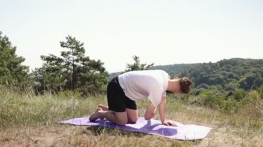 Yoga odağı, huzurlu doğa, otlak alanı. Doğanın huzuru ile çevrili beyaz tişörtlü ve siyah şortlu genç adam çimenli çayırlarda mor hasırda yoga yapıyor..