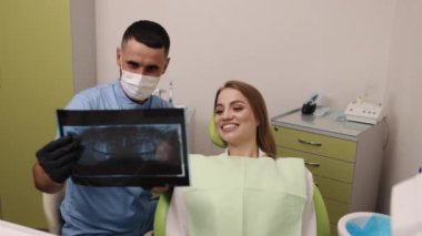 Diş maskesi, diş prosedürü, diş donanımı. Diş sandalyesindeki hasta mavi önlüklü ve maskeli dişçi röntgenini gösterirken gülümsüyor..