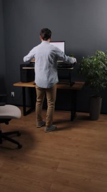 Çalışma istasyonu kurulumu, ayakta masa, ergonomik masa. Ahşap döşeme ve ergonomik sandalyeli modern ofiste, çiçeklerle süslenmiş kapüşonlu genç adam ayakta duran masa, dikey video.