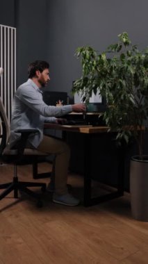 Kahveni yudumla, iş adamı, odaklanmış ifade. Açık mavi gömlekli bir adam, bitkilerle çevrili şık bir ofiste masasını kullanırken kahve içer. Dikey