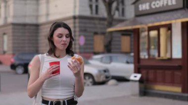 Çiğneme burger, açık hava yemeği, kırmızı bardak. Kot pantolonlu ve beyaz bluzlu genç kadın hamburger yiyor ve kahve standının dışında kırmızı kahve bardağı tutuyor..