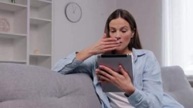 Şok olmuş kadın, Tablet sahnesi, ev zarafeti. Tablete bakarken ağzını kapatan, modern oturma odasındaki gri koltukta oturan şaşkın kadın..