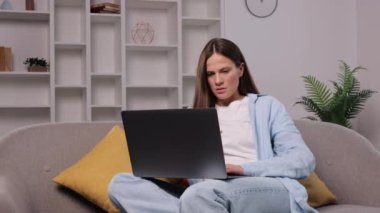 Boyun ağrısı, çalışma gerginliği, kanepe oturması. Genç bir kadın kanepede oturuyor, dizüstü bilgisayarla çalışırken boynunu tutuyor, gergin ya da kötü duruşu gösteriyor..
