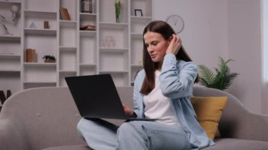 El sallama, dizüstü bilgisayar sohbeti, neşeli ekran. Kanepede oturmuş, gülümseyen ve dizüstü bilgisayarına el sallayan neşeli genç bir kadın..