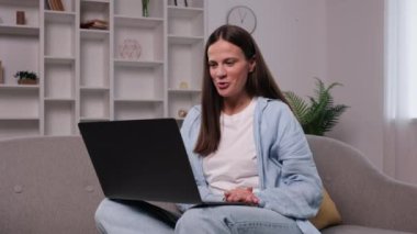 Video çağrısı, laptop ev, çapraz bacaklı kadın. Neşeli genç bir kadın evdeki kanepede bağdaş kurup oturuyor ve dizüstü bilgisayarındaki video çağrısına katılarak gülüyor..
