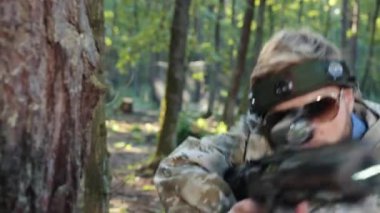 İnsan kamuflajı, ağaç duruşu, düşman savaşı. Kafasında işaret olan kamuflaj elbiseli bir adam ağaca tutuldu. Titizlikle lazer etiketli tabancayla düşmana ateş etti.
