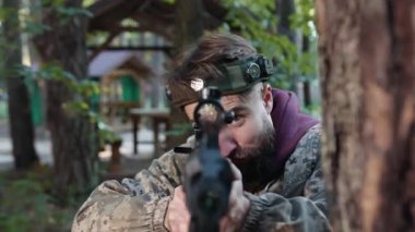 Taktik teçhizat, düşman çatışması, orman çatışması. Spor kamuflaj teçhizatı ve kafasında işaretler olan adam ağaca dayanıyor, lazer tabancasını nişan alıyor ve ağaçlık alanda düşmanı vuruyor..