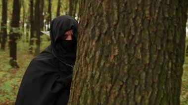 Boyalı el, gölgeli figür, gizemli figür. Gölgeli ormanda, boyalı ellerle kapüşonlu bir figür ağaç gövdesini kavrıyor, gizlilik ve belirsizlik uyandırıyor..
