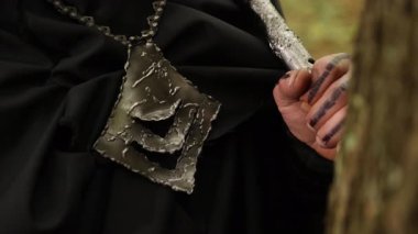 Sert mücevherler, silah sapı, siyah pelerin. Yakından bakınca zincirden sarkıtılmış engebeli metal bir kolye ve siyah pelerinlilere karşı elle kavrayan bir silah görülüyor..