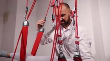 Fizik tedavi uzmanı, klinik destek, işlevsel yardım. Kadın hasta tedavi masasında yatıyor, kırmızı terapi kemeriyle askıda, beyaz önlüklü fizik terapistin de yardımıyla..