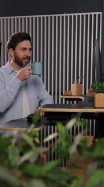 Adam kahve içiyor, kahve içiyor, iş adamı. Açık mavi gömlekli bir adam modern ofiste yeşillikle süslenmiş bir şekilde masasında çalışırken kahvesini yudumluyor. Dikey