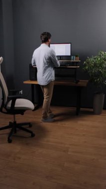 Daimi bir iş, masa başı, masa kurulumu. Monitörle ayakta duran, açık mavi gömlekli adam modern ofiste ahşap döşeme ve bitkilerle çalışıyor, dikey video..