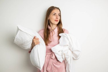 Sabahlık giymiş genç bir kadın beyaz bir arka planda yastık ve çarşaf çekiyor..