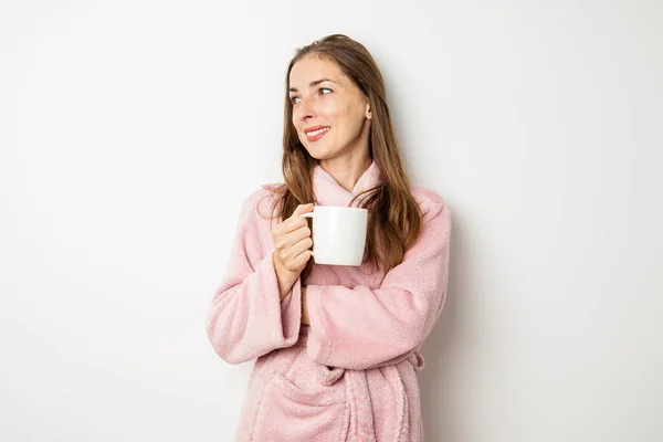 Smiling young woman in a bathrobe with a cup on a white background.