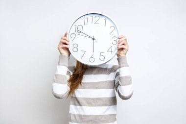Kadınların elleri beyaz bir duvar saati tutuyor. Yüzler arka planda görünmüyor. 