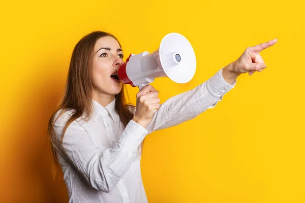 Young woman in a white shirt makes an announcement in a megaphone and points a finger on a yellow background. Hiring concept, help wanted. Banner.