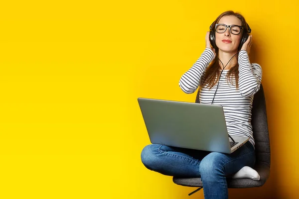 stock image Young woman in headphones listening to music and sitting on chair and holding laptop on isolated yellow background.