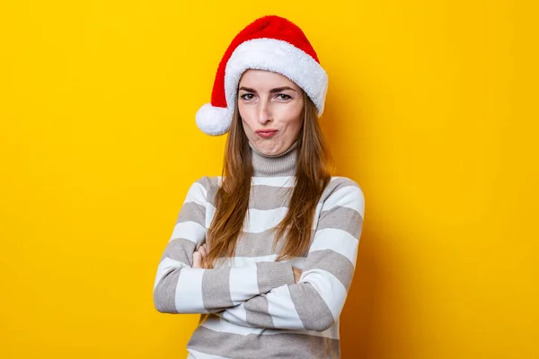 Frowning young woman in Santa Claus hat on a yellow background.