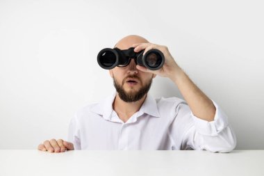 Genç adam beyaz bir arka planda beyaz bir masanın altından dışarı bakan dürbünle bakar..