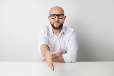 Genç adam beyaz bir arka planda masada otururken elini uzatır..