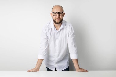 Gözlüklü genç adam beyaz arka planda bir masada duruyor..