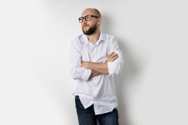 Beyaz tişörtlü genç kel adam kollarını beyaz bir arka planda kavuşturmuş duruyor..