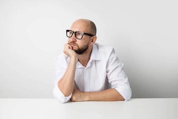 stock image Young man holds his hand under his chin while sitting at a table on a white background.