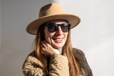 Güneş gözlüklü ceketli, beyaz bir duvara yaslanmış gülümseyen genç bir kadın.