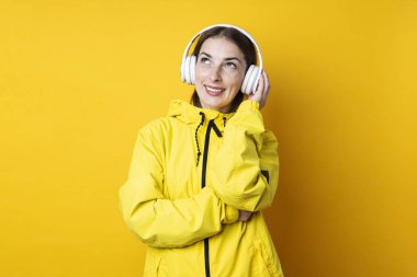 Sarı ceketli, kulaklıklı, gülümseyen genç bir kadın sarı bir arka plana bakıyor.